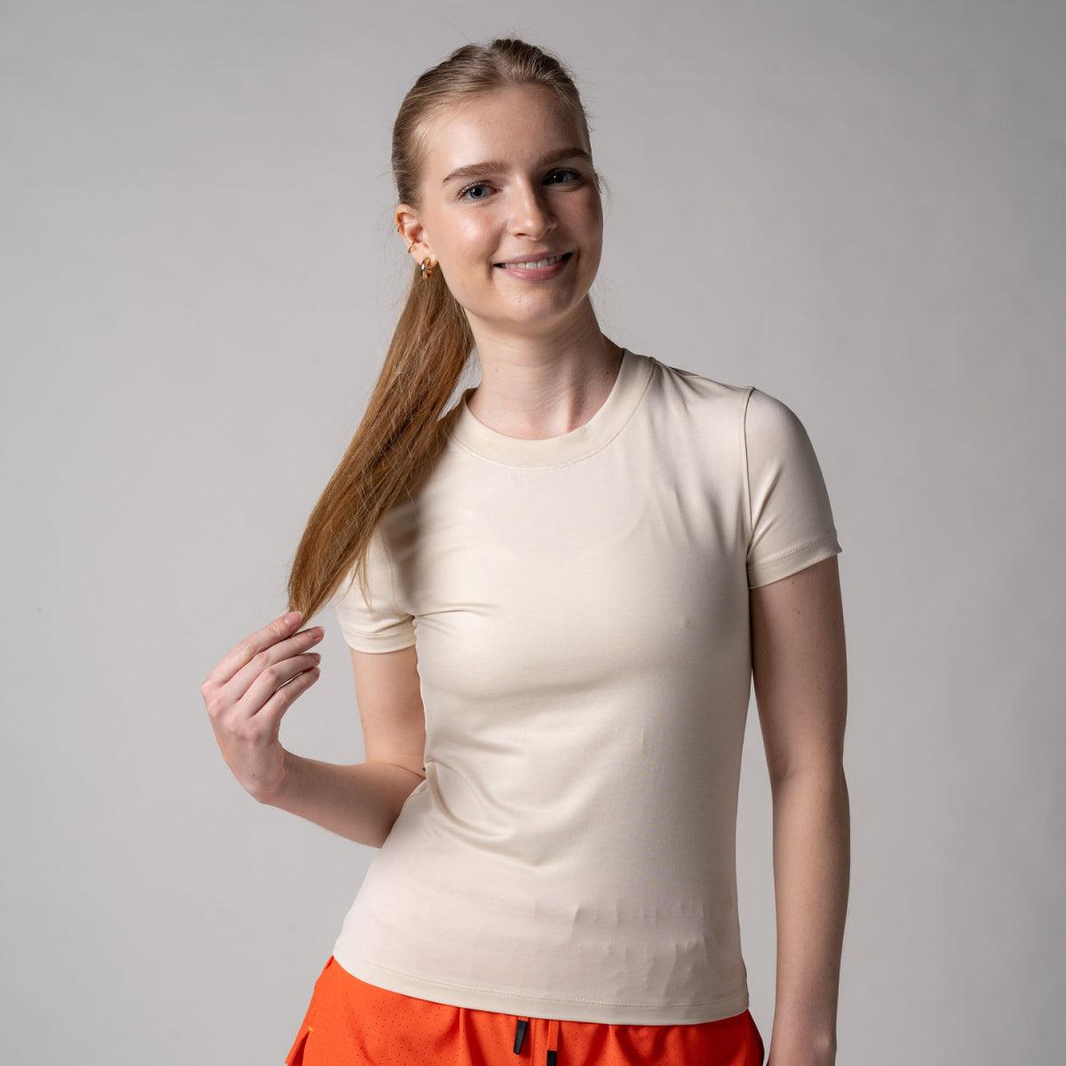Frau trägt beige, eng anliegendes T-Shirt mit kurzen Ärmeln und orangefarbener Hose, lächelt und hält eine Haarsträhne.