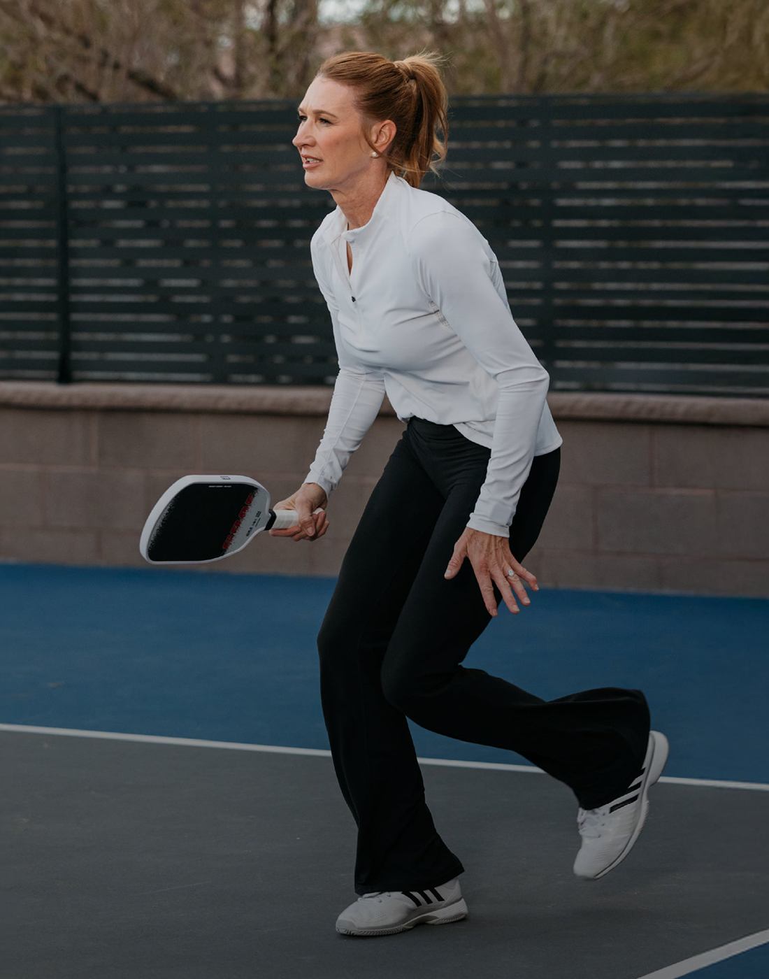 Frau in weißem Langarmshirt und schwarzer Hose spielt Pickleball auf Außenplatz mit Schläger in der Hand.