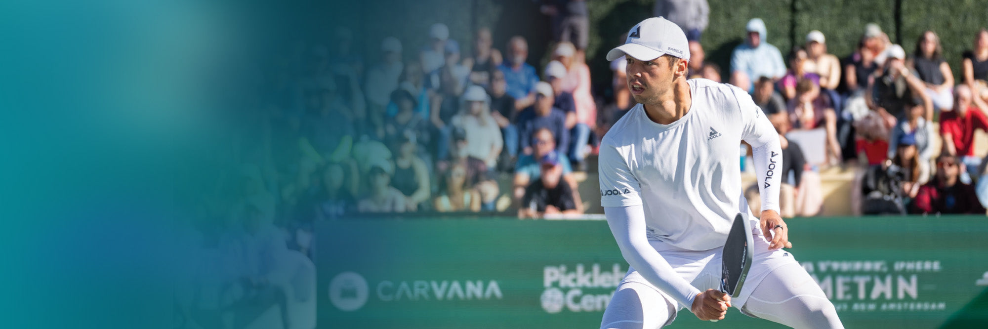 Mann in weißer Sportkleidung spielt Pickleball vor Publikum auf Outdoor-Platz, hält Schläger bereit zum Schlag.