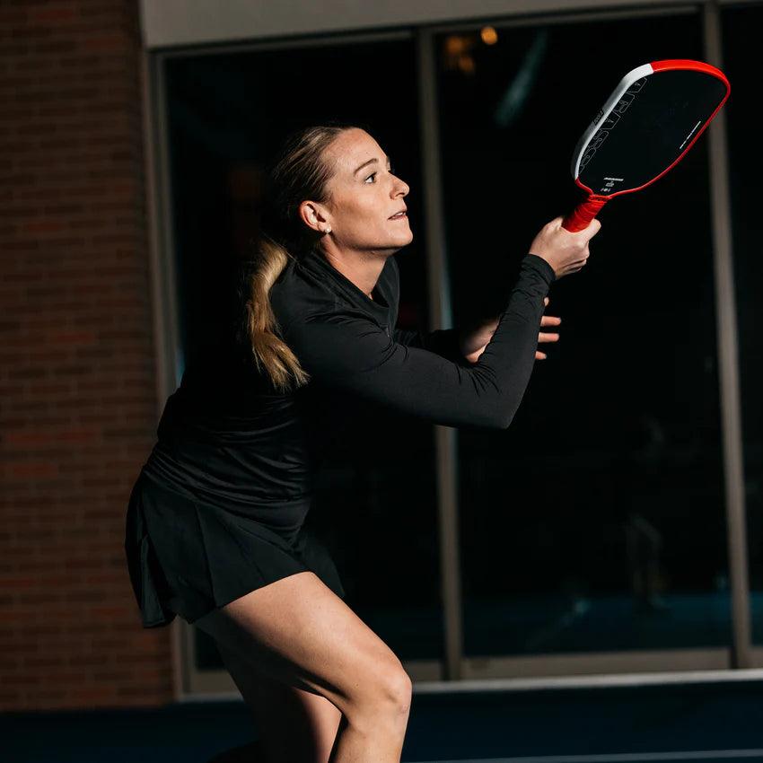 Frau in schwarzer Sportkleidung spielt Pickleball mit rotem Schläger in Hallenumgebung.